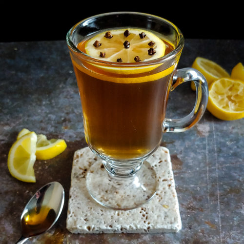 warm cocktail in a glass with a lemon wheel garnish, spoon and lemons
