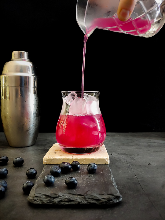 pouring a magenta tequila blueberry cocktail into an ice-filled glass