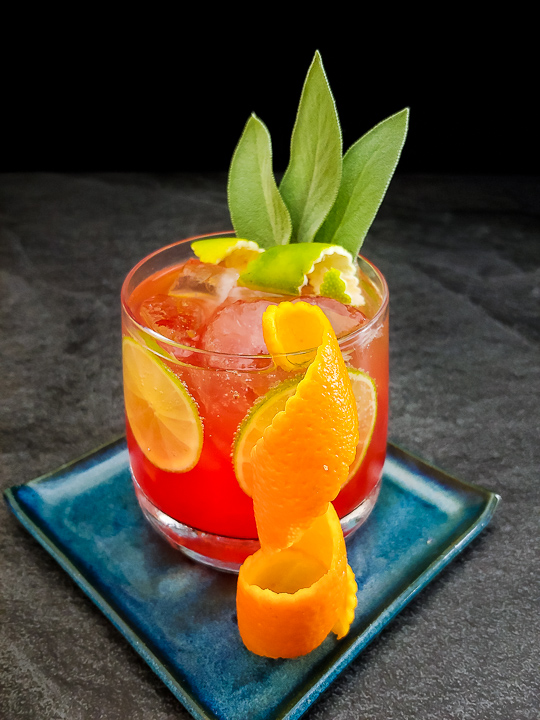 red cocktail in a rocks glass with sage leaf, orange and lime zest garnish