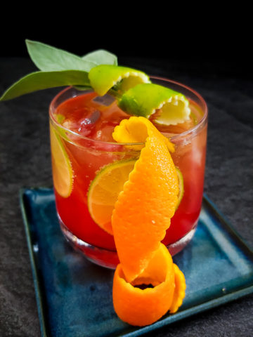red cocktail in a rocks glass with sage leaf, orange and lime zest garnish
