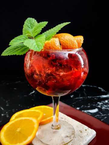 Bitter Bourbon Spritz red cocktail in a wine glass with orange and mint garnish