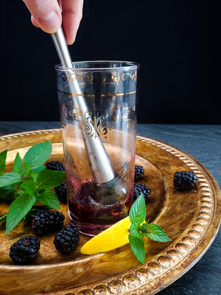 muddling blackberry bourbon cocktail in highball glass with lemon and mint garnish