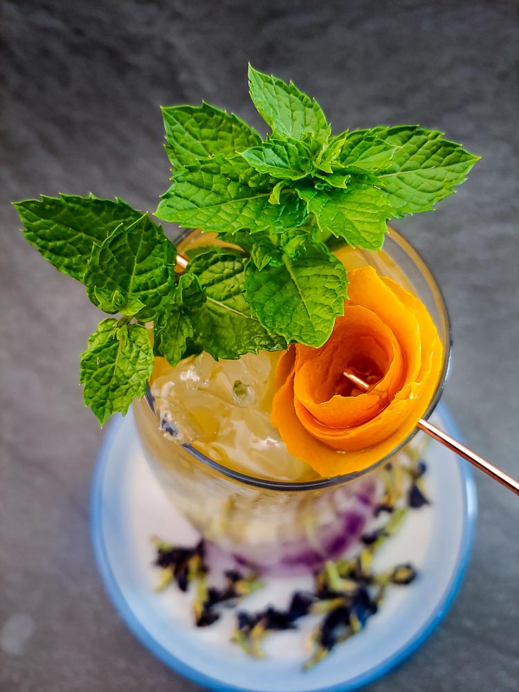 Bourbon cooler in tall glass with purple, white and yellow layer, orange rose and mint garnish