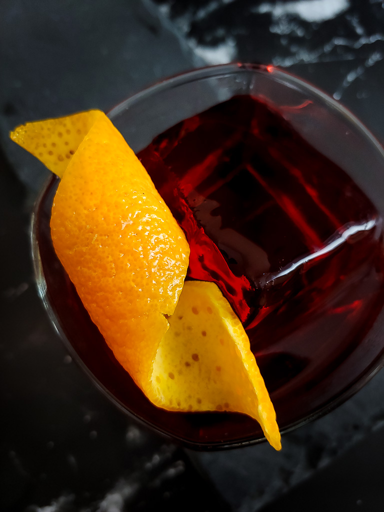 red cocktail over ice in a glass with orange peel garnish.