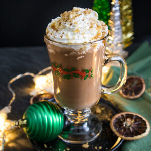 Caramel Orange Hot Chocolate topped with whipped cream and caramel drizzle on a plate with christmas lights and ornaments