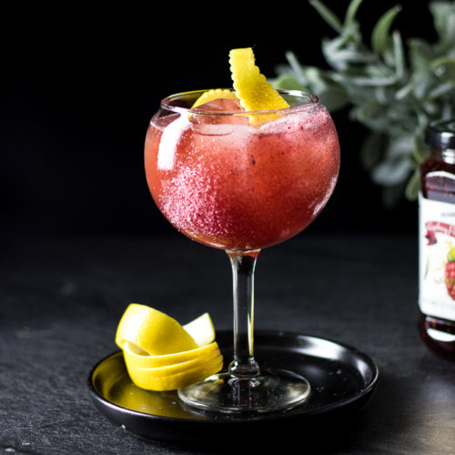 Raspberry Rue Jam Cocktail in a wine glass with ice, garnished with a lemon twist