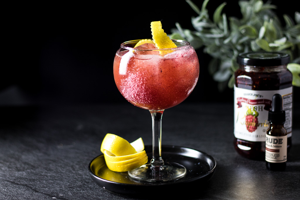 Raspberry Rue Jam Cocktail in a wine glass with ice, garnished with a lemon twist
