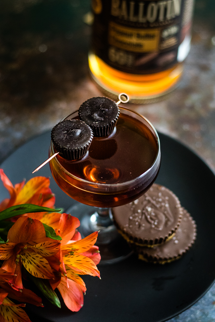 Peanut Butter Chocolate Cocktail in a coupe glass with two peanut butter cups, flowers and bottle of ballotin whiskey
