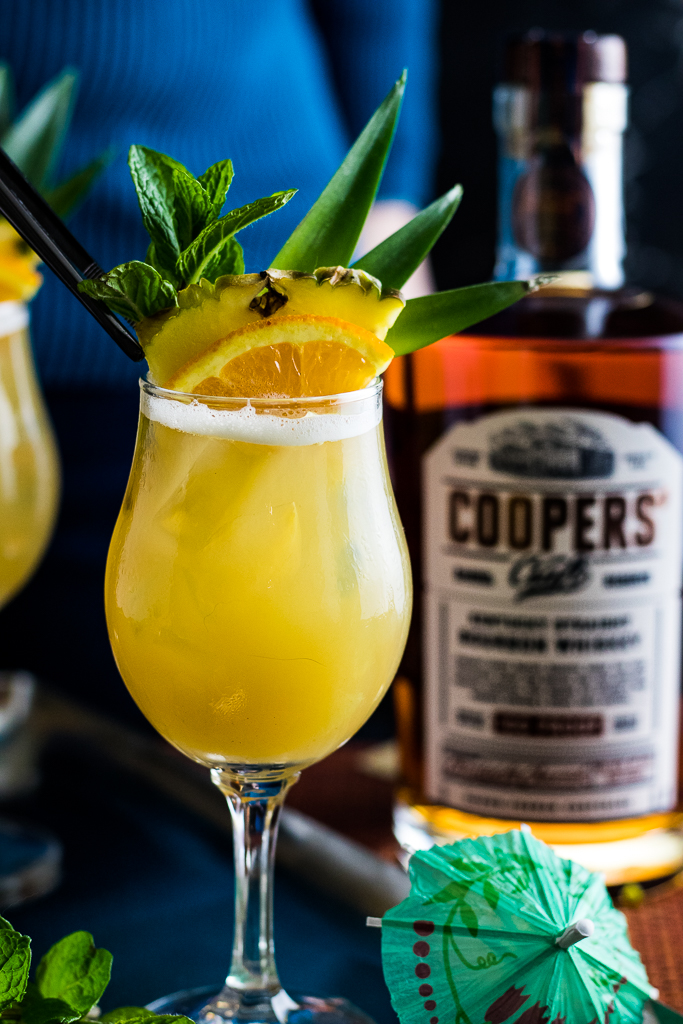 Whiskey Tiki Sour in two wine glasses with pineapple spear garnish, pineapple, orange and mint