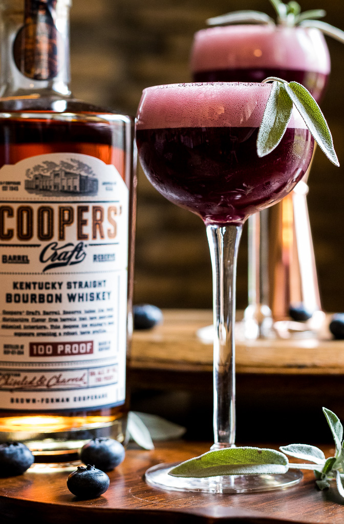 Coopers' Craft Cocktail - Blueberry Sour with sage leaf garnish and a bottle of Coopers' Craft on a gold tray