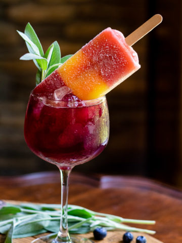 blueberry bourbon smash cocktail in tall wine glass with ice and topped with a sorbet popsicle and garnished with fresh sage