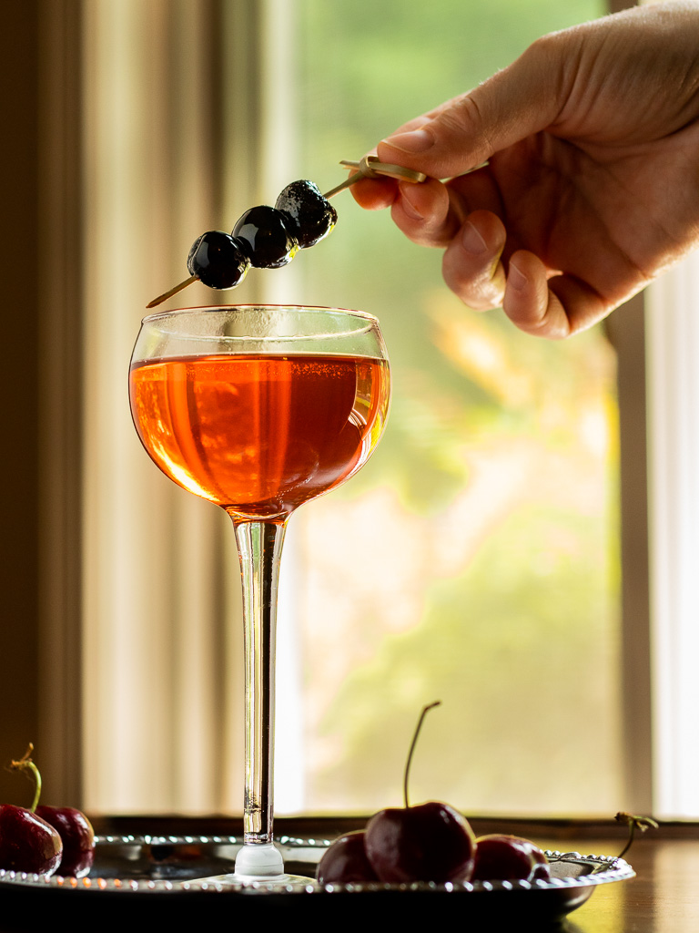 Aperol bourbon cocktail in a coupe glass with garnish of sweet cherries