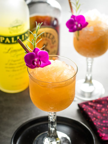 Lavender Lemon Bourbon Slushies - two in coupe glasses with lavender and flower garnish and bottles of whiskey and limoncello