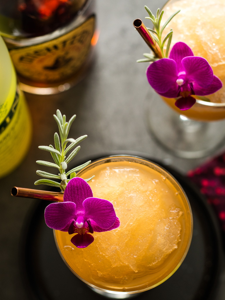 Lavender Lemon Bourbon Slushies - two in coupe glasses with lavender and flower garnish and bottles of whiskey and limoncello