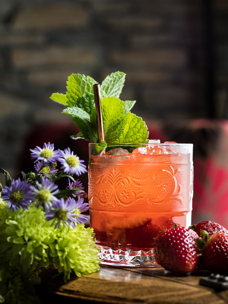 Strawberry whiskey smash in a rocks glass on a wooden tray with flowers and strawberries
