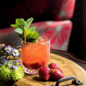 Strawberry whiskey smash in a rocks glass on a wooden tray with flowers and strawberries