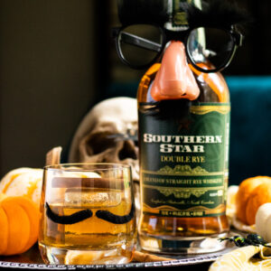 Halloween Whiskey Cocktail in a glass with a mustache, and fake eyeglasses on a bottle of rye. Pumpkins and skulls in the background