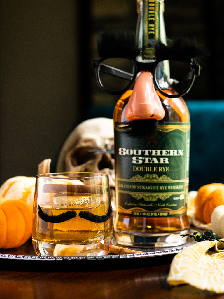 Halloween Whiskey Cocktail in a glass with a mustache, and fake eyeglasses on a bottle of rye. Pumpkins and skulls in the background
