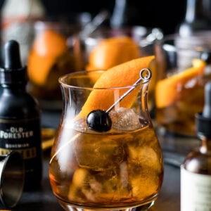 a perfect old fashioned in a rocks glass over ice with bitters and other old fashioned surrounding it. Garnished with orange peel and cherry