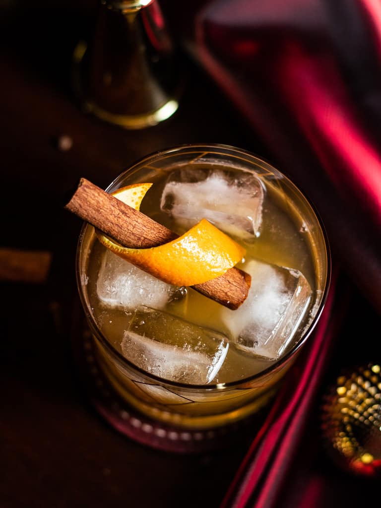 Christmas Old Fashioned in a rocks glass with an orange and cinnamon stick garnish, surrounded by red fabric and gold ornaments.