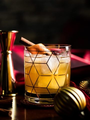 Christmas Old Fashioned in a rocks glass with an orange and cinnamon stick garnish, surrounded by red fabric and gold ornaments.