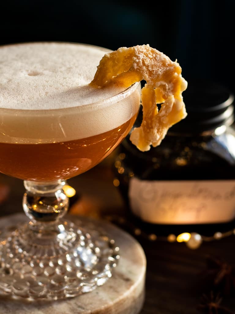 gingerbread whiskey sour in a coupe glass with a candied ginger garnish on a tray