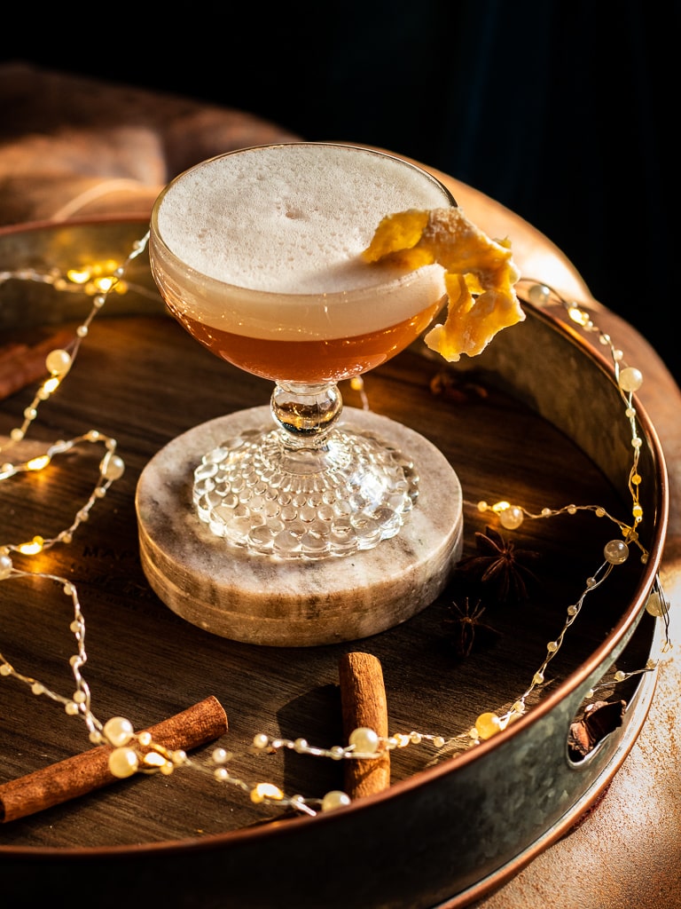 gingerbread whiskey sour in a coupe glass with a candied ginger garnish on a tray