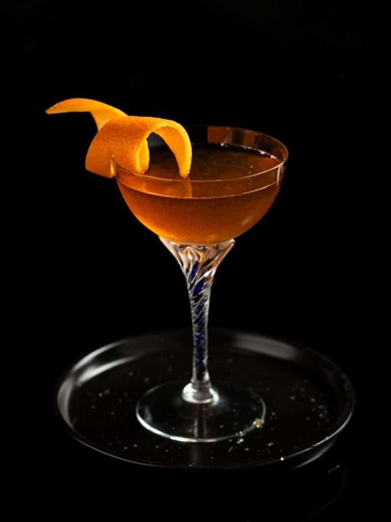 Orange marmalade Breakfast manhattan in a coupe class with an orange twist garnish on a small back plate against a black background