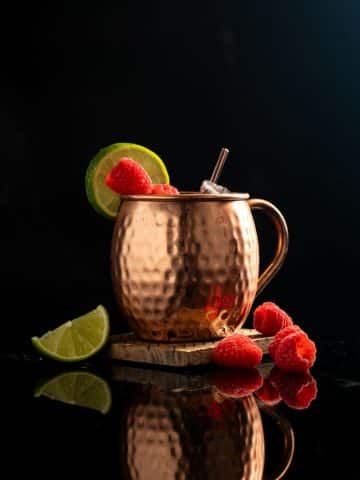 Raspberry Mule cocktail in a copper mug with raspberry garnish and lemon wheel against a black background