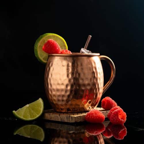 Raspberry Mule cocktail in a copper mug with raspberry garnish and lemon wheel against a black background