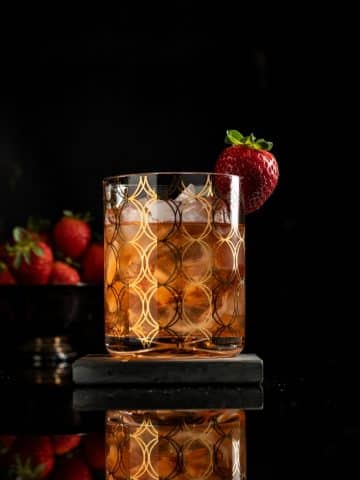 strawberry cardamom old fashioned in a rocks glass with ice and a strawberry garnish against a black background