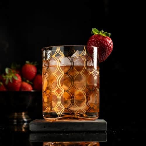 strawberry cardamom old fashioned in a rocks glass with ice and a strawberry garnish against a black background