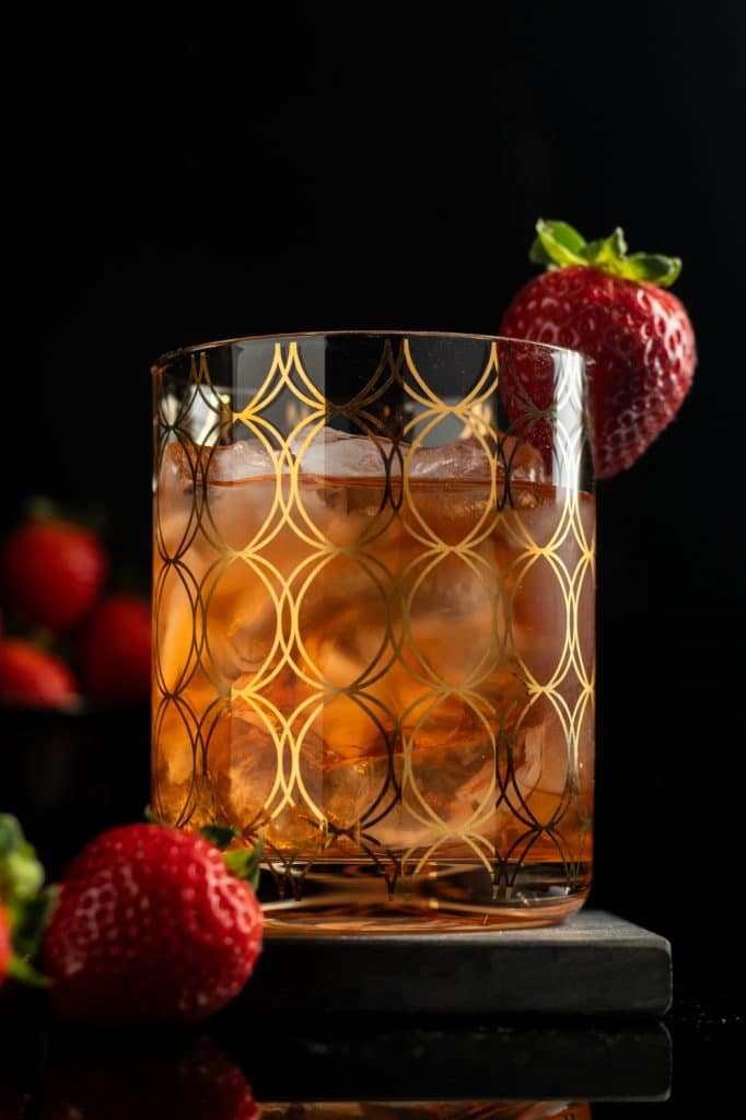 closeup strawberry cardamom old fashioned in a rocks glass with ice and a strawberry garnish against a black background