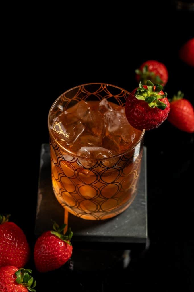 strawberry cardamom old fashioned in a rocks glass with ice and a strawberry garnish against a black background